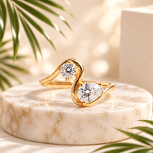 Load image into Gallery viewer, A gold ring with two pear-cut clear gemstones in a swan-like curved design, displayed on a round white marble pedestal with soft-focus palm leaves and a beige background.